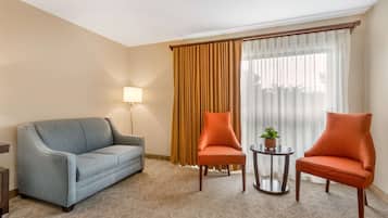 A hotel room with a light blue sofa, two orange armchairs, a small round table with a plant, a lamp, and a large window with curtains.