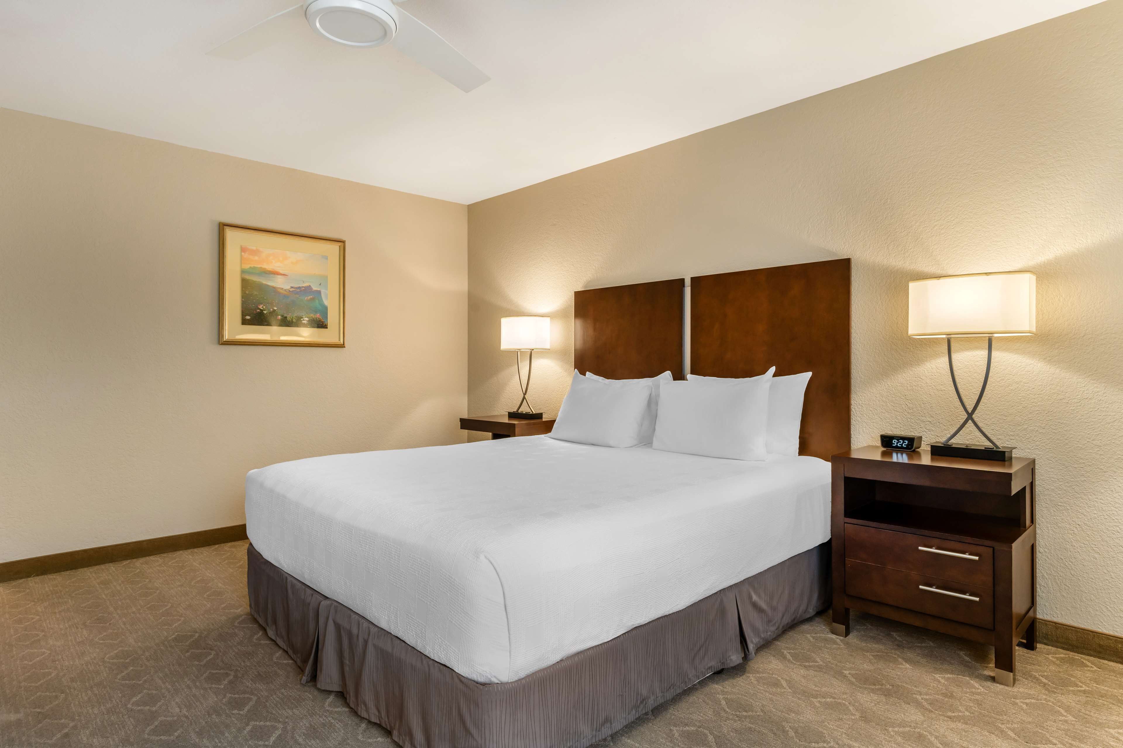 A hotel room with a large bed, two bedside tables, a ceiling fan, a wall-mounted painting, and a nightstand with a lamp.