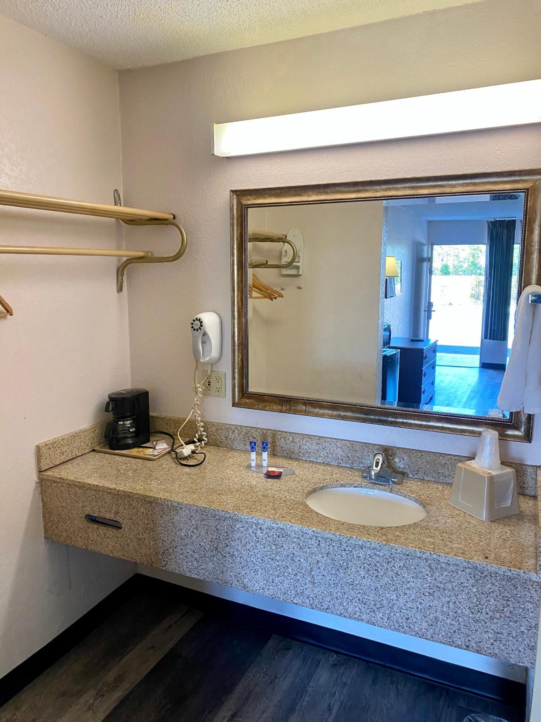A hotel bathroom with a granite countertop, a sink, a mirror, a towel rack, a hairdryer, and a coffee maker.