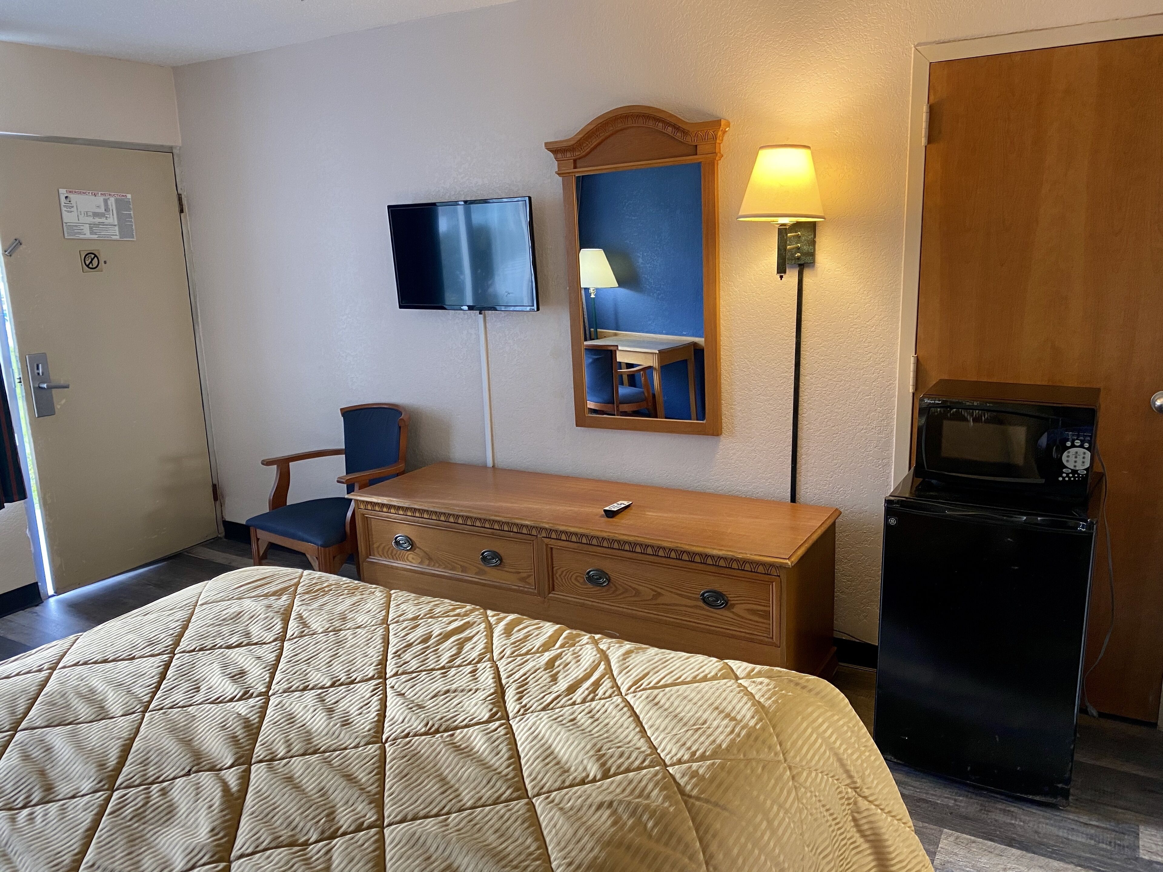 A hotel room with a bed, a wooden dresser, a desk with a chair, a television, a mirror, a lamp, and a microwave.