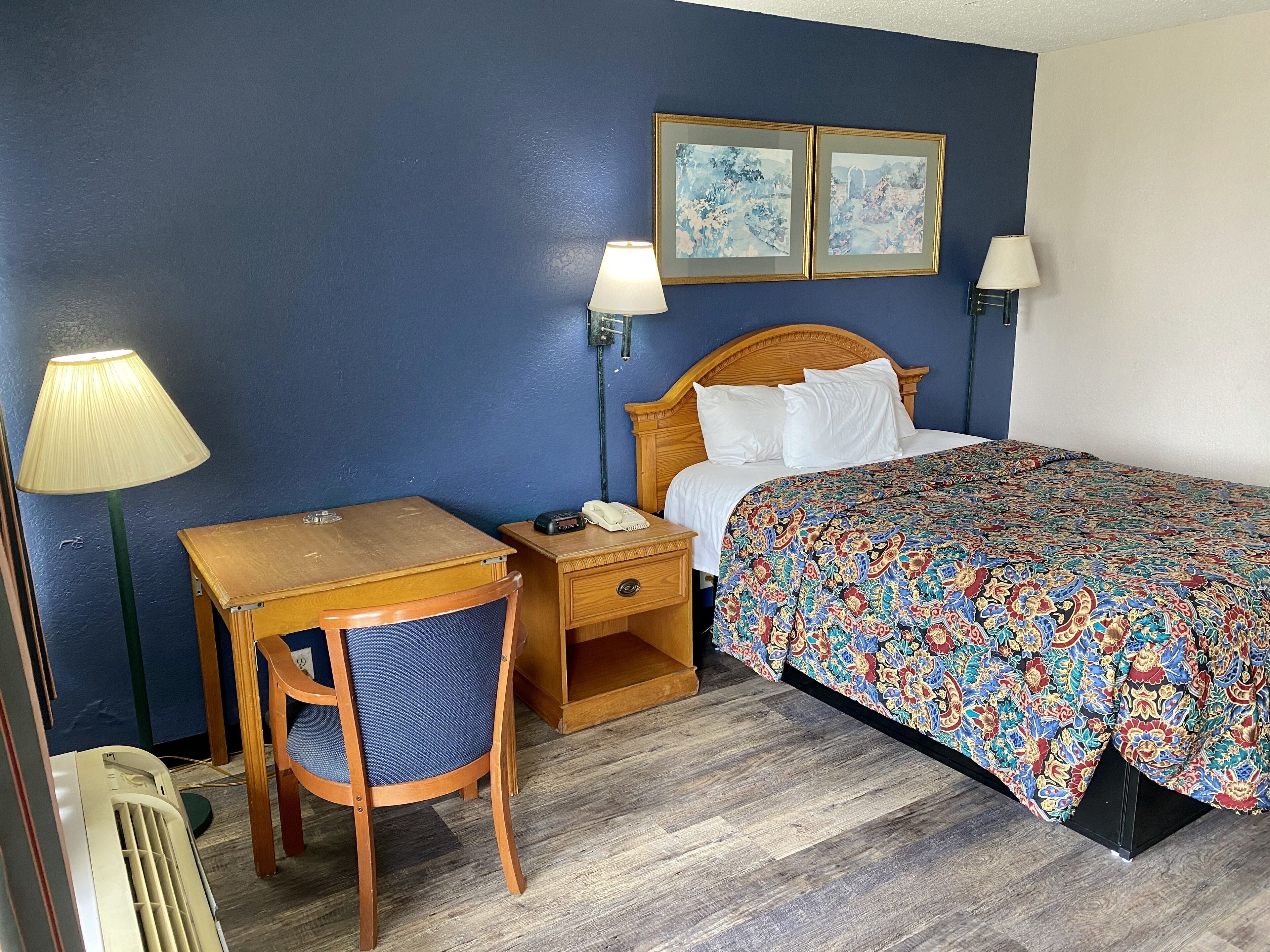 A hotel room with a wooden desk, chair, and a bed with a colorful patterned bedspread. There are two wall-mounted lamps and two framed pictures above the bed.