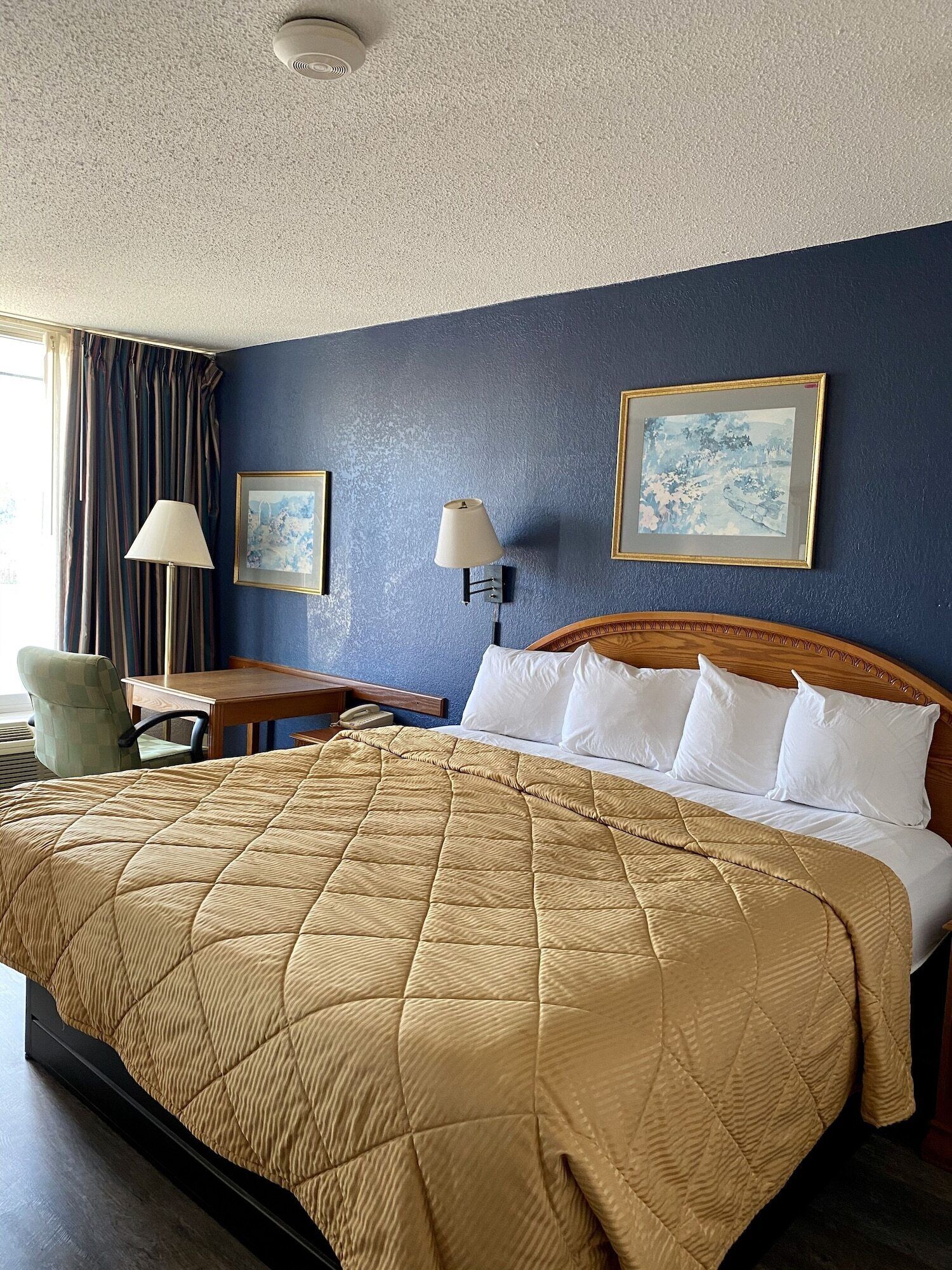 A hotel room with a large bed, two wall-mounted lamps, a wooden headboard, a desk with a chair, and two framed pictures on the wall.