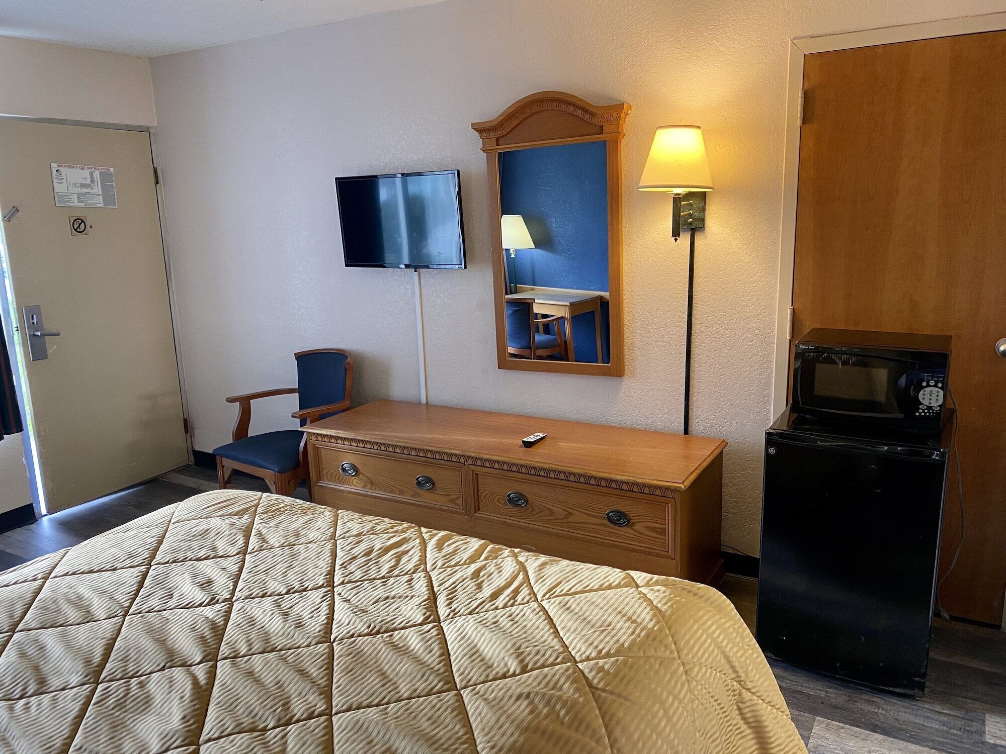 Een hotelkamer met een bed, een houten dressoir, een bureau met stoel, een televisie, een spiegel, een lamp en een magnetron.