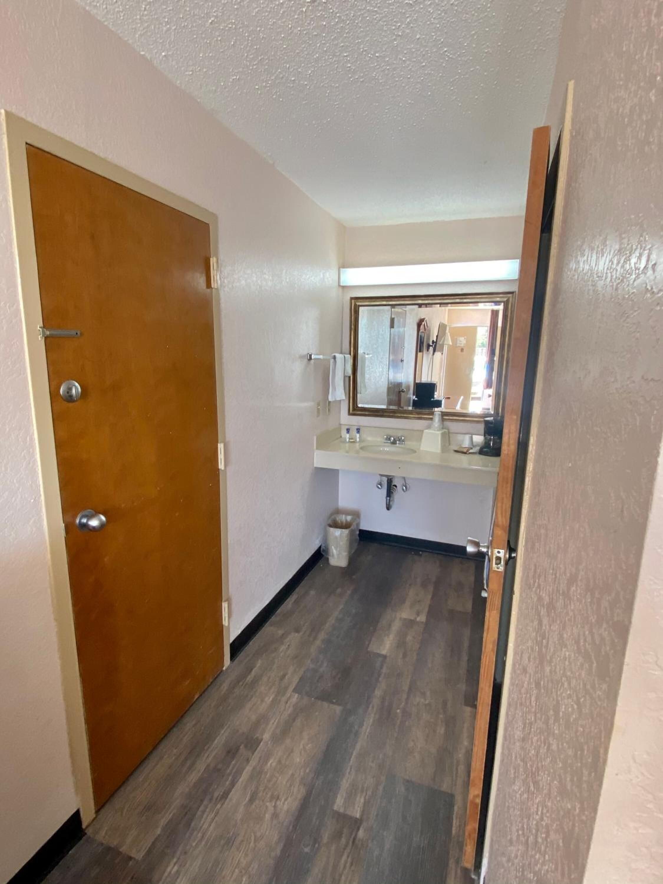 A hallway with a wooden door, a bathroom with a sink and mirror, and a trash can.