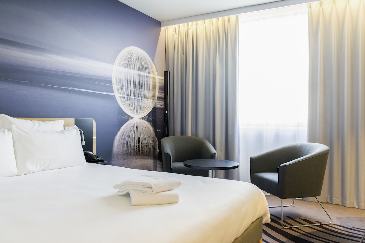A hotel room with a large bed, two chairs, a small table, and a wall art piece.
