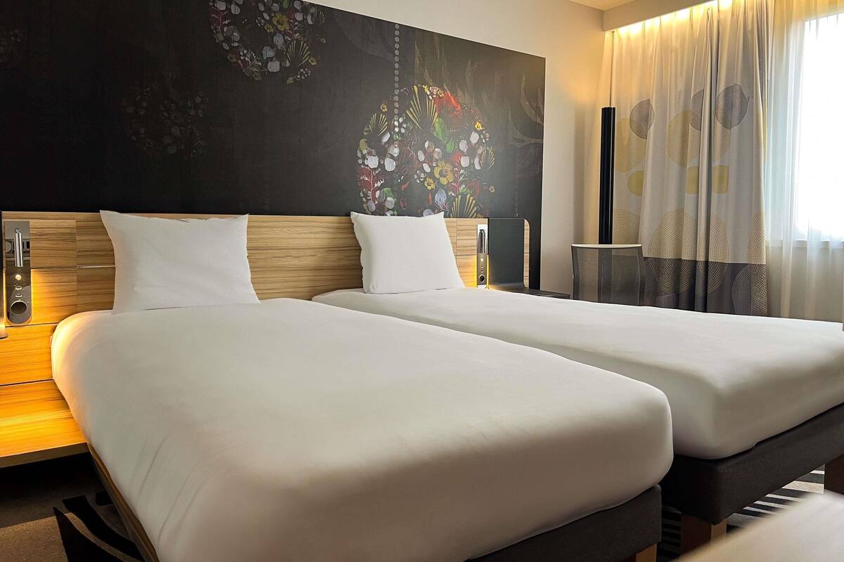 A hotel room with two beds, a dark wall with floral artwork, a bedside table with a lamp, and sheer curtains.
