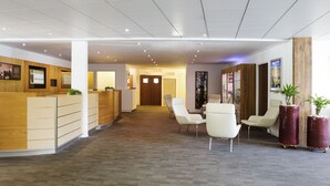 A reception area with a wooden reception desk, white chairs, and a carpeted floor. - Novotel Limoges Le Lac (Limoges)