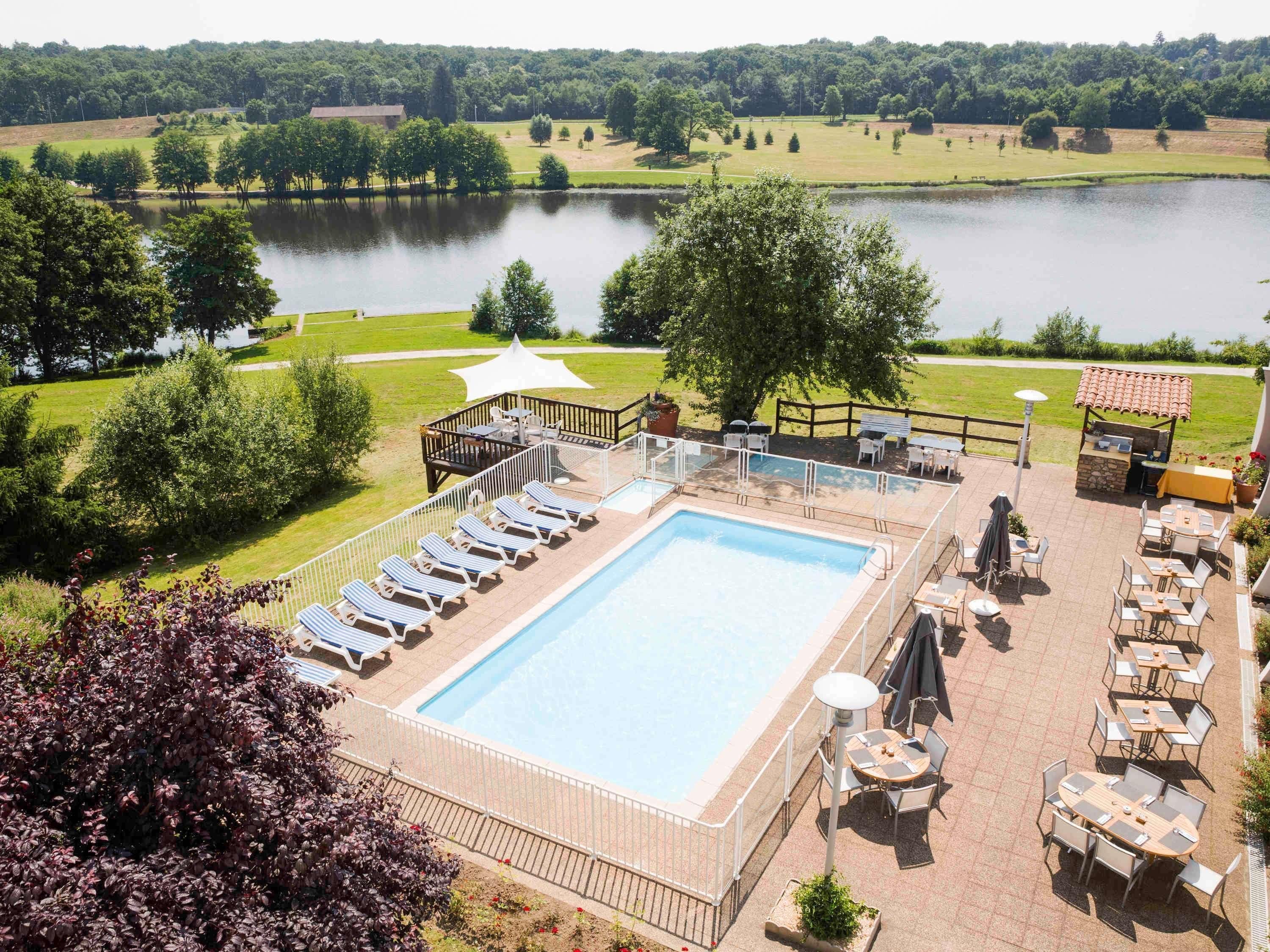 Ein rechteckiger Pool mit Liegestühlen und Tischen, umgeben von einer Terrasse mit Blick auf einen See und Bäume.
