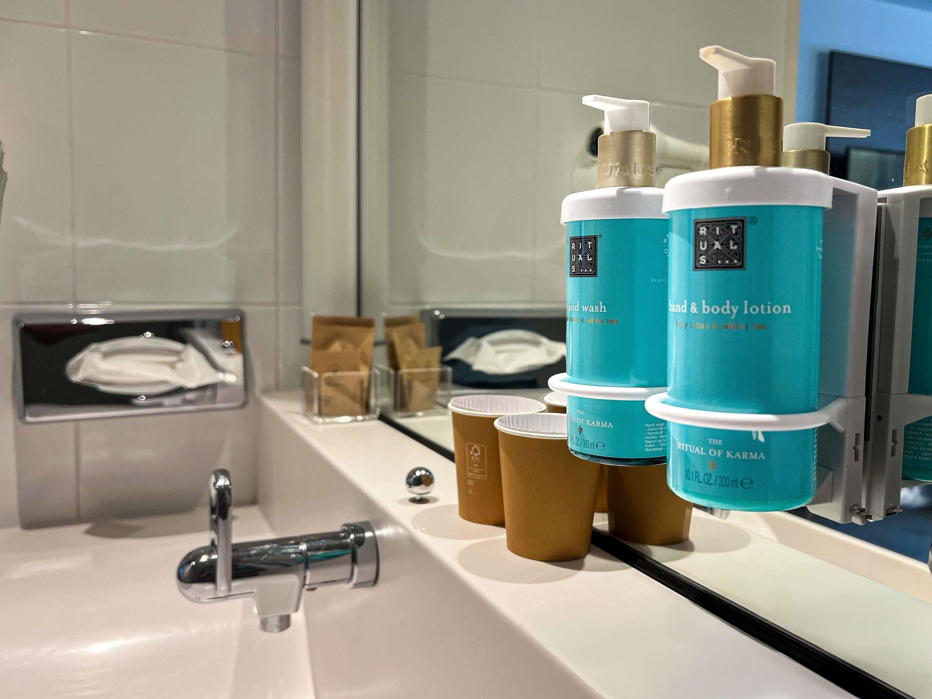 A bathroom with a sink, a mirror, and various toiletries including hand wash and body lotion bottles.