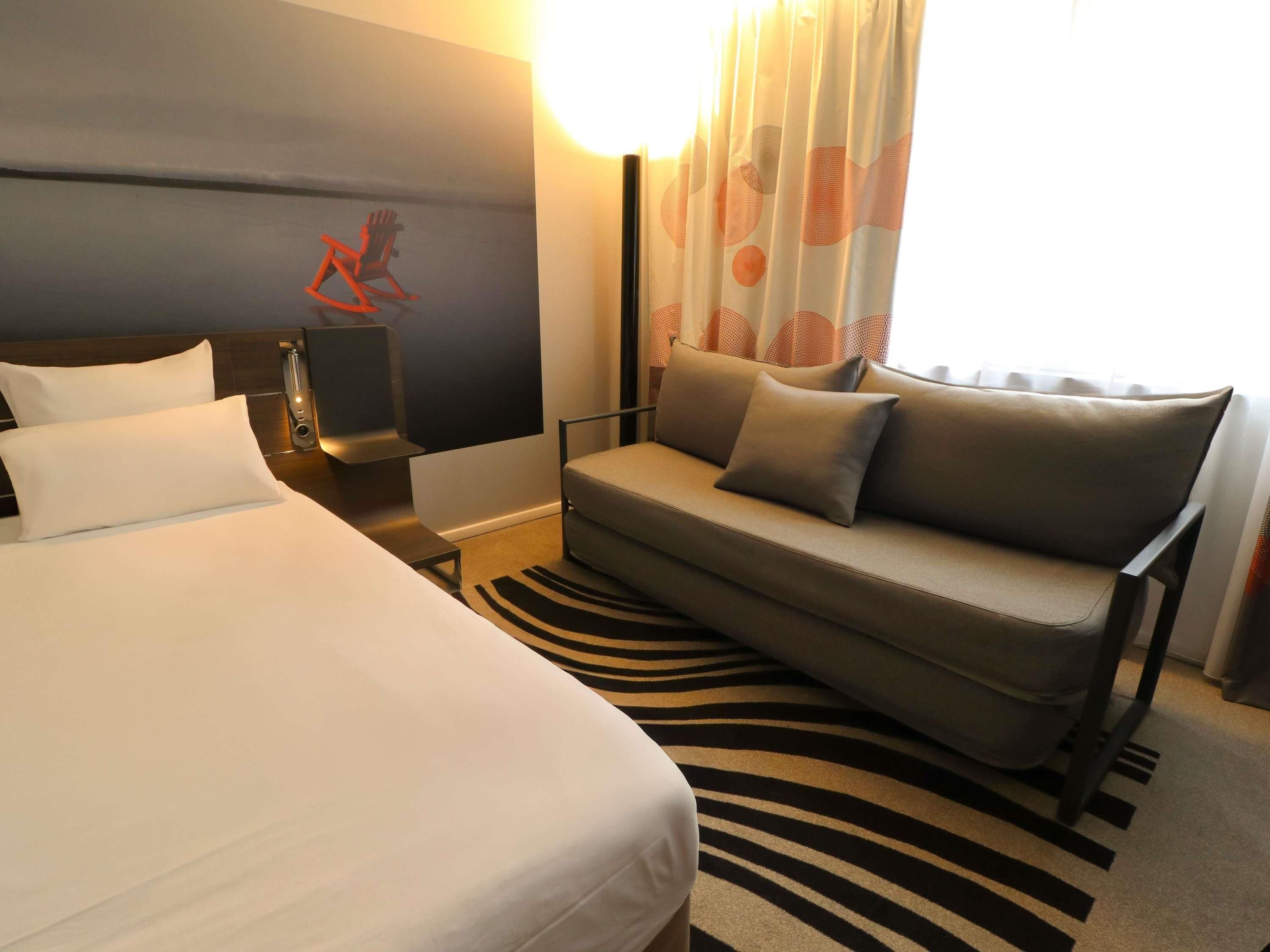 A hotel room with a bed, a sofa, a nightstand, and a wall-mounted TV.