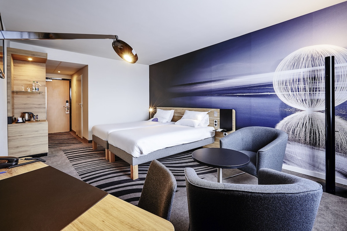 A modern hotel room with a large bed, a desk, and a chair. There is a painting on the wall and a lamp on the ceiling.