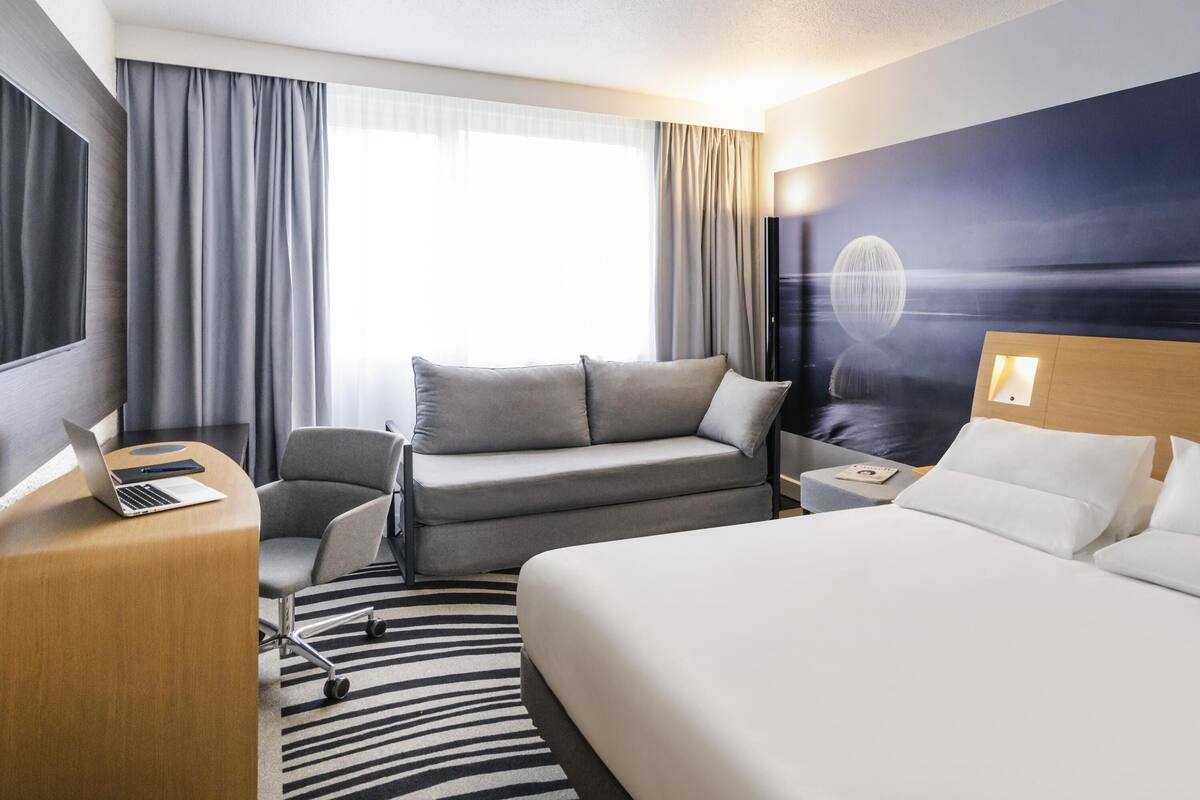 A hotel room with a bed, a sofa, a desk with a laptop, a nightstand, and a wall-mounted TV.