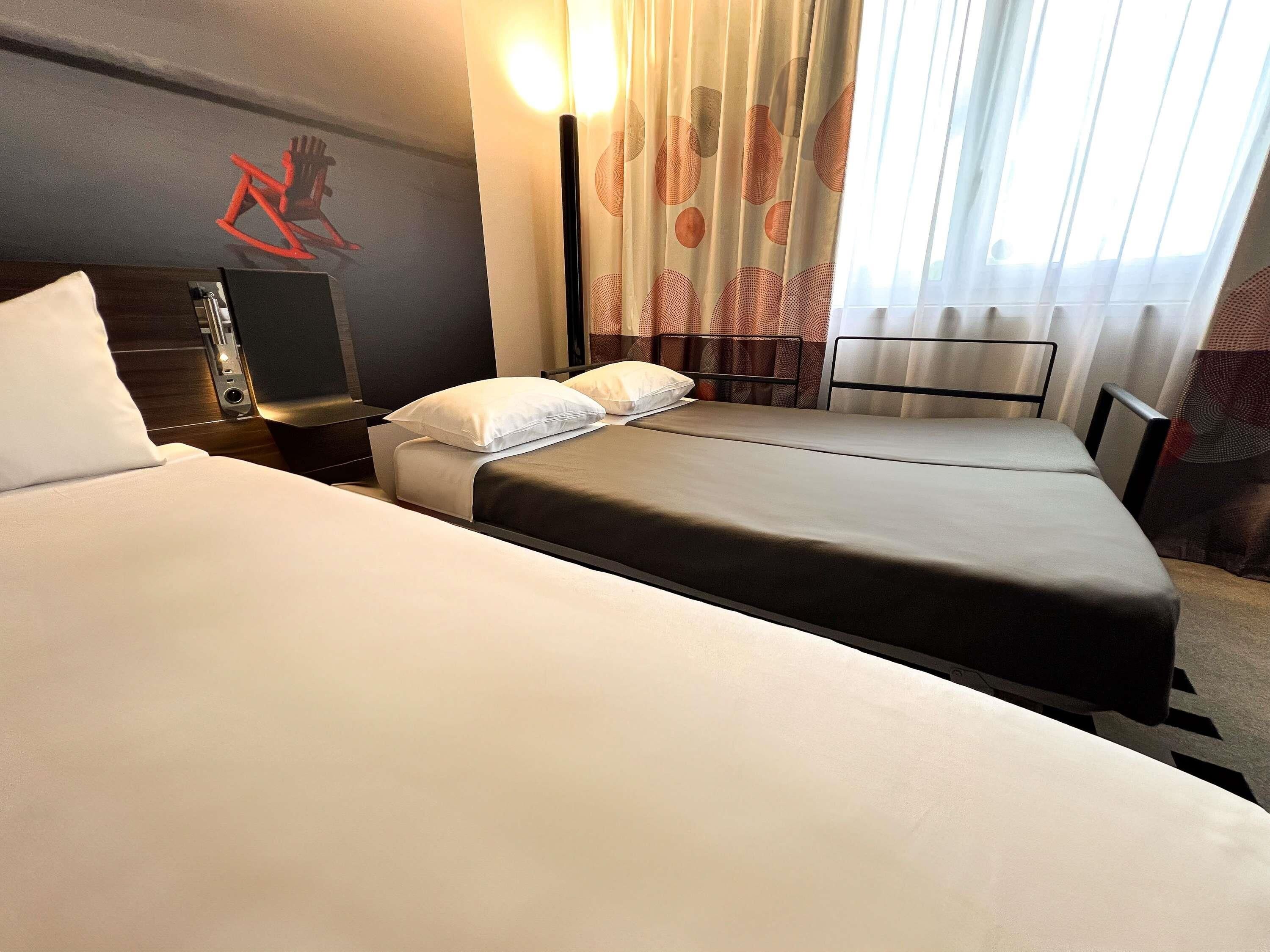 A hotel room with two beds, a nightstand, a lamp, and a wall-mounted artwork.