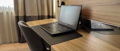 Una laptop sobre un escritorio de madera con una silla, cortinas y un televisor en la pared.