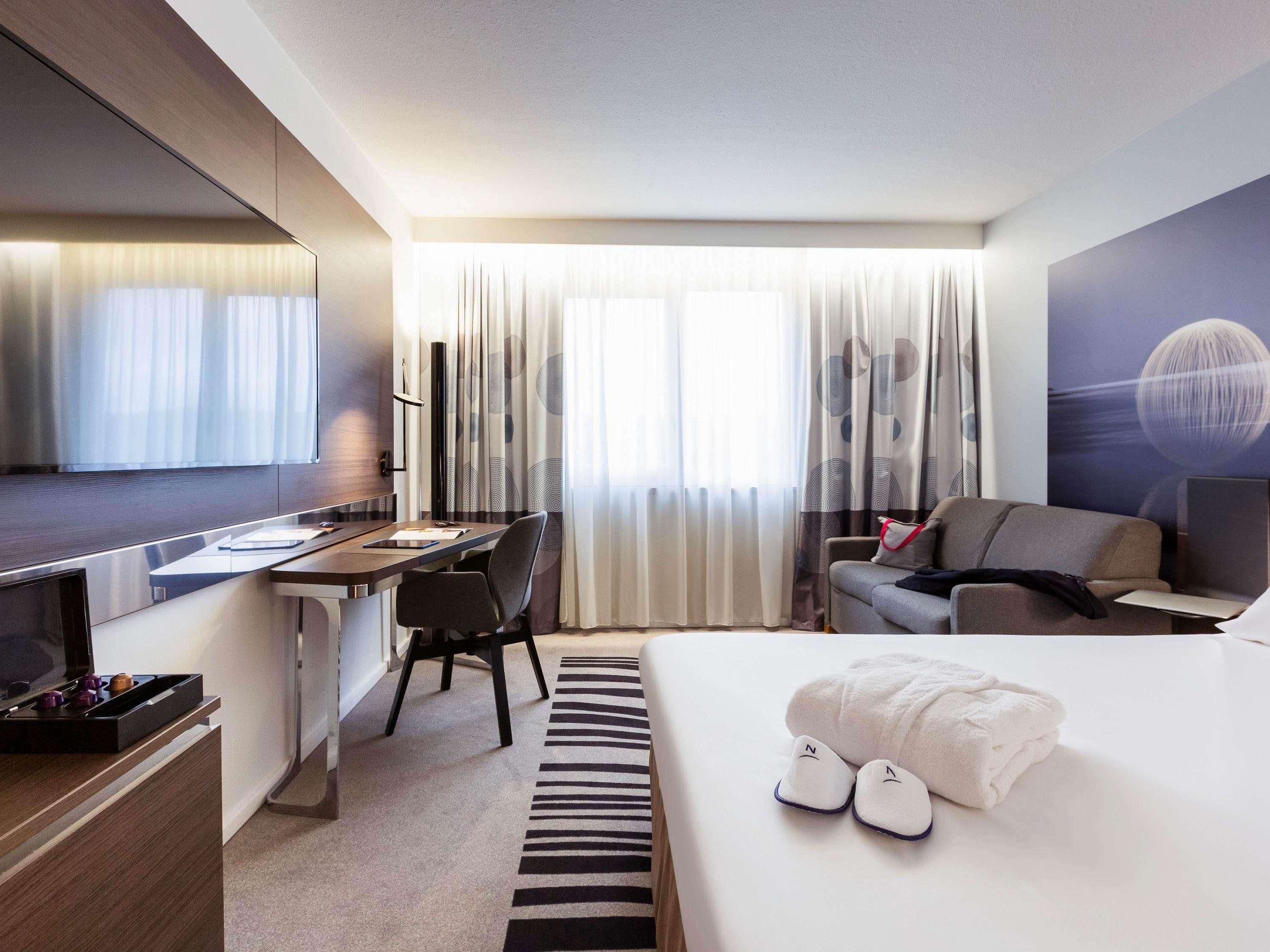 Camera d'albergo con un letto, una scrivania, una sedia, una televisione e un'ampia finestra con tende.