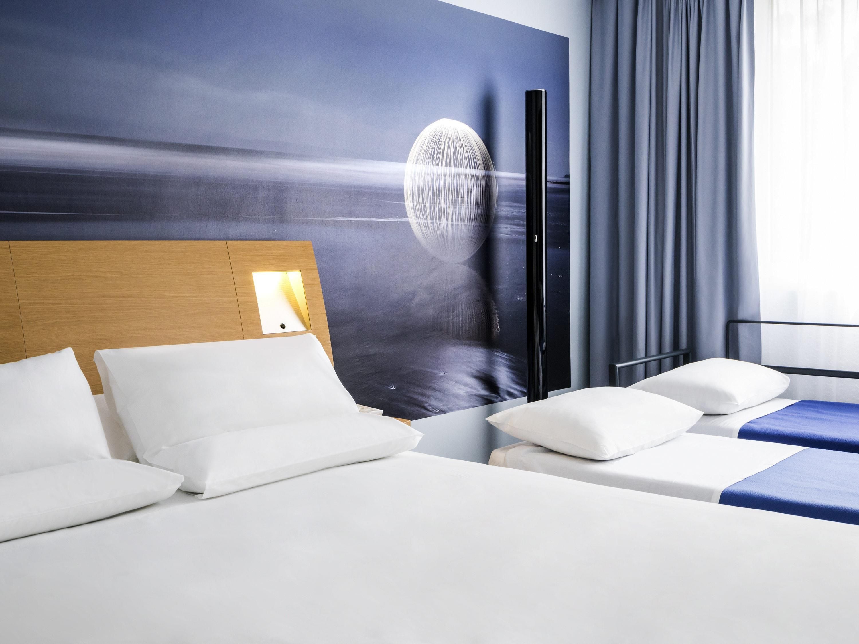 A hotel room with a large wall mural, a wooden headboard, and a bed with white bedding.