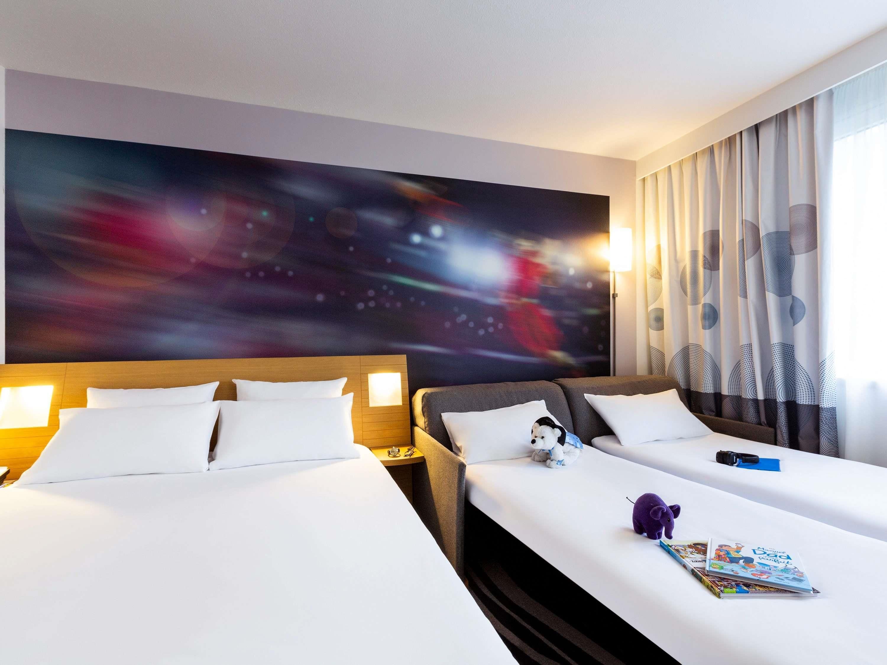 A hotel room with two beds, a large abstract painting on the wall, a nightstand with a lamp, and a window with curtains.