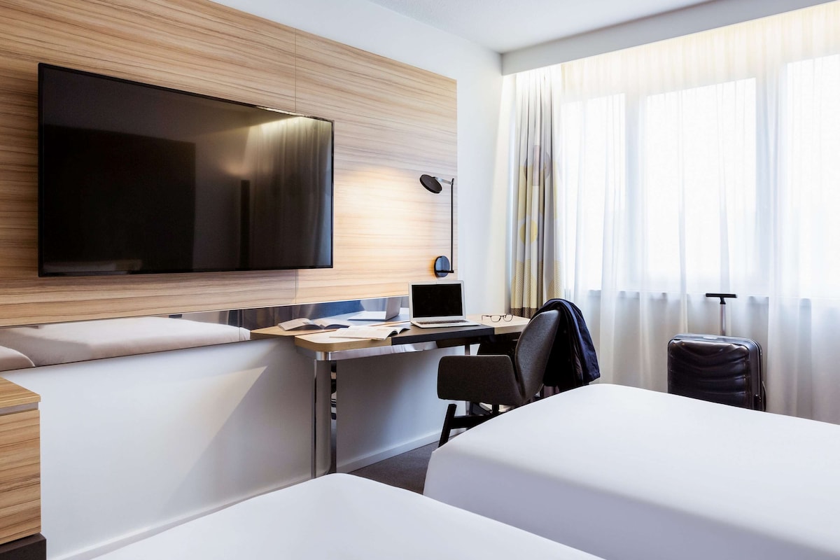 A modern hotel room with a large flat-screen TV, a desk with a laptop, a chair, and a bed.