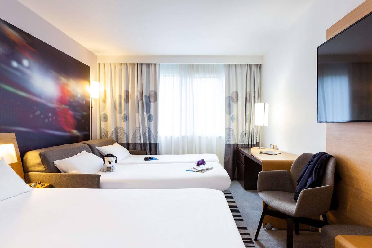 A hotel room with two beds, a desk, a chair, a TV, and a large window with curtains.