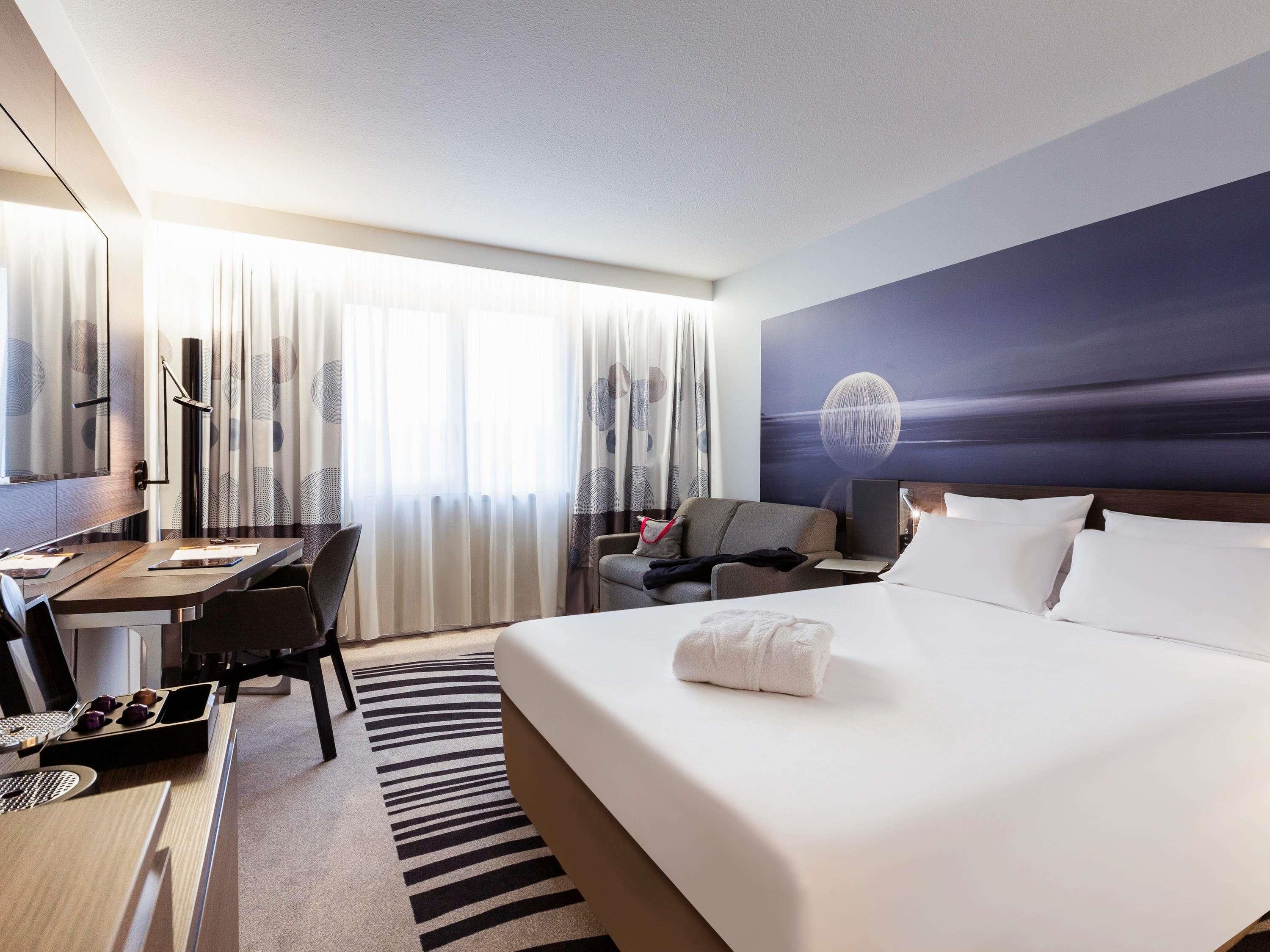 A hotel room with a bed, a desk with a laptop, a chair, a sofa, and a large window with curtains.