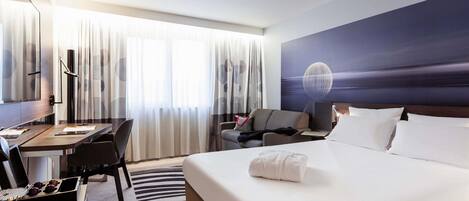 Habitación de hotel con cama, escritorio con una computadora portátil, una silla, un sofá y un ventanal con cortinas.