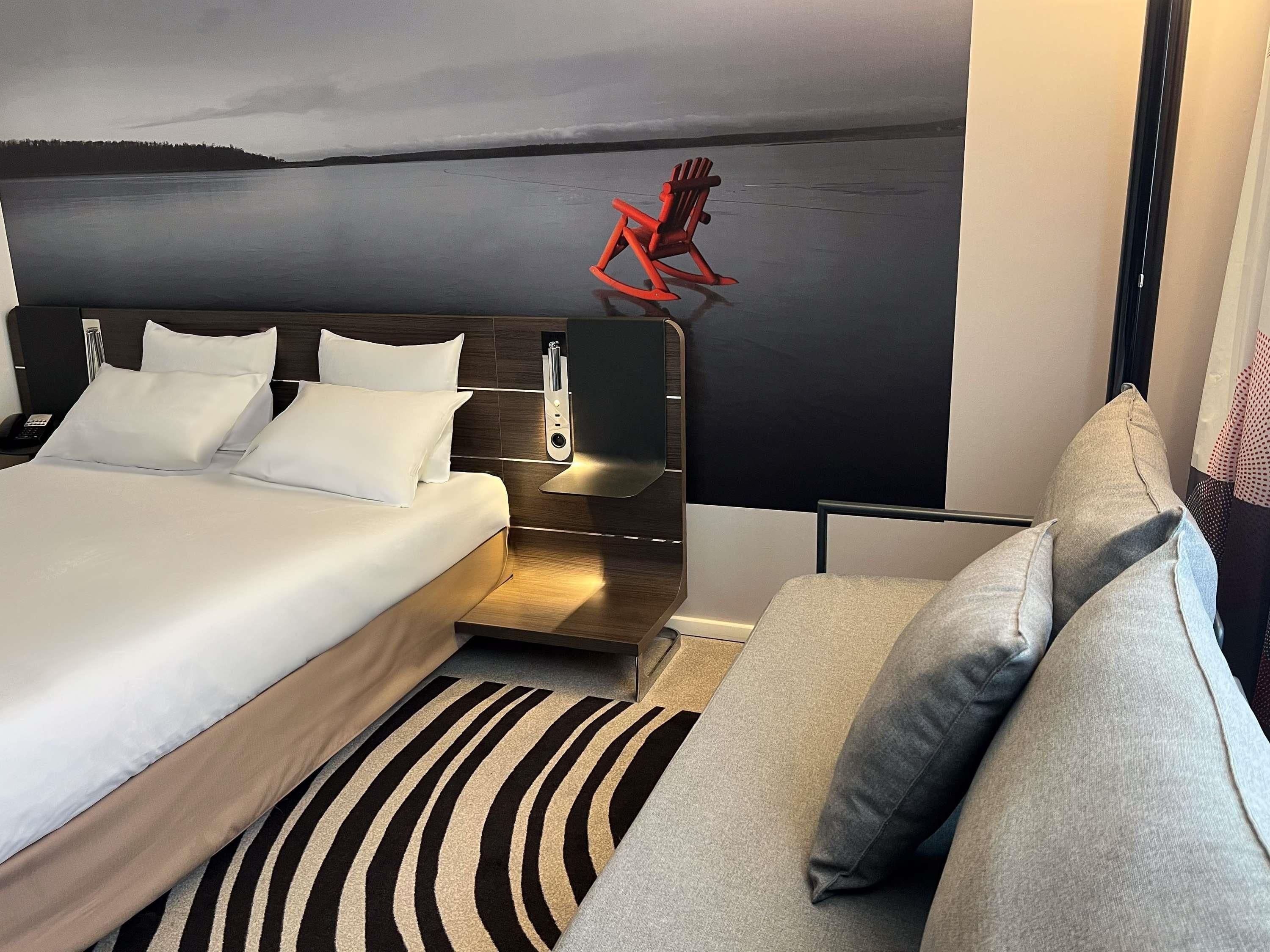 A modern hotel room with a large bed, a sofa, a wall-mounted TV, and a view of a lake.