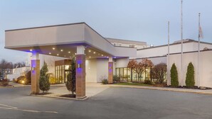 Una entrada de hotel moderna con un corredor cubierto, fachada de vidrio y un área ajardinada con arbustos y árboles.