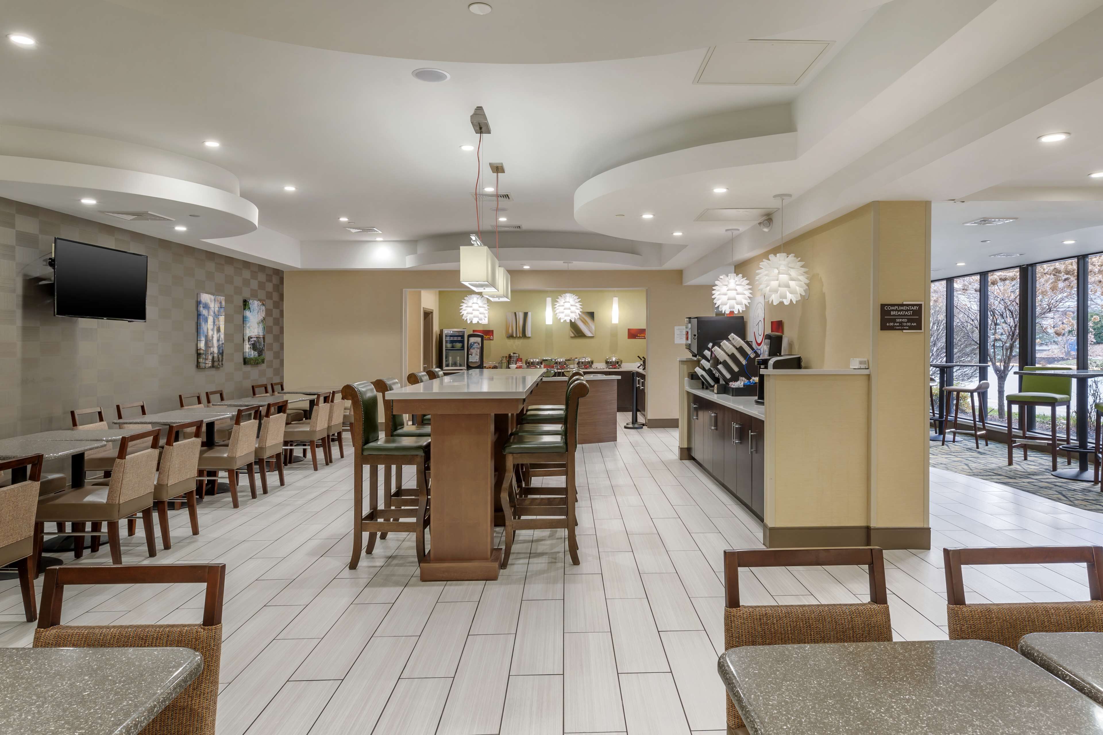 A spacious hotel lobby with multiple dining tables and chairs, a central bar area, and a large window providing a view of the outdoors.