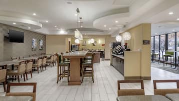A spacious hotel lobby with multiple dining tables and chairs, a central bar area, and a large window providing a view of the outdoors.