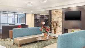 Un moderno hall de hotel con un área de descanso que incluye sofás tapizados en azul, una mesa de centro con un jarrón de flores y un televisor de pantalla plana grande montado en una pared oscura.