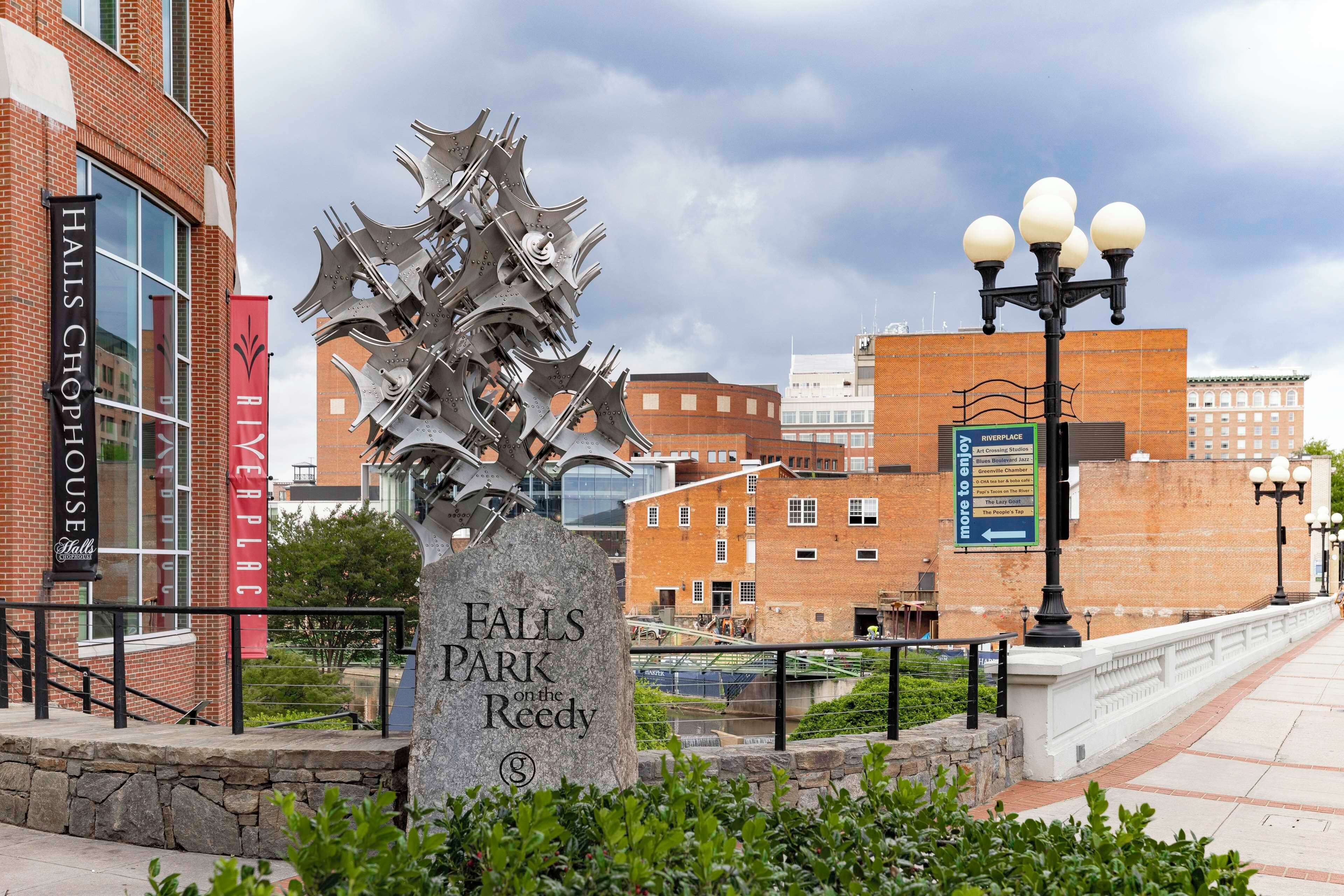 A stone monument with the inscription "Falls Park on the Reedy" surrounded by greenery, with a cityscape in the background including a brick building and a sign for "Halls Chop House."