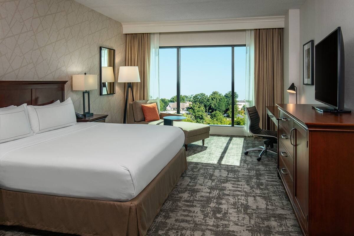 Une chambre d’hôtel avec un grand lit, un bureau avec une télévision, une chaise et une vue sur la verdure par la fenêtre.