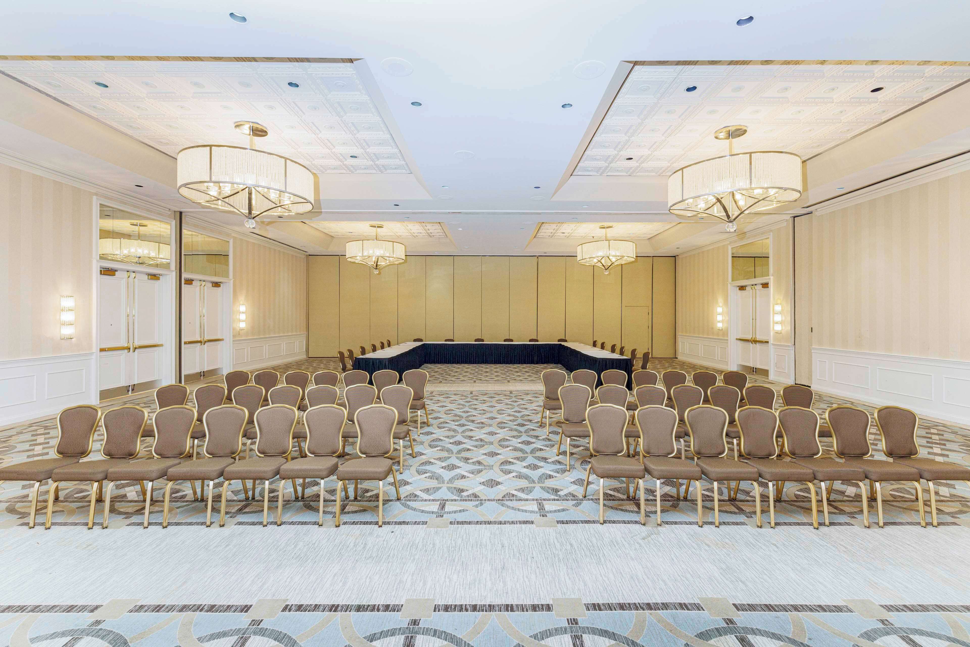 A conference room with a long table and rows of chairs, carpeted floor, and chandeliers.