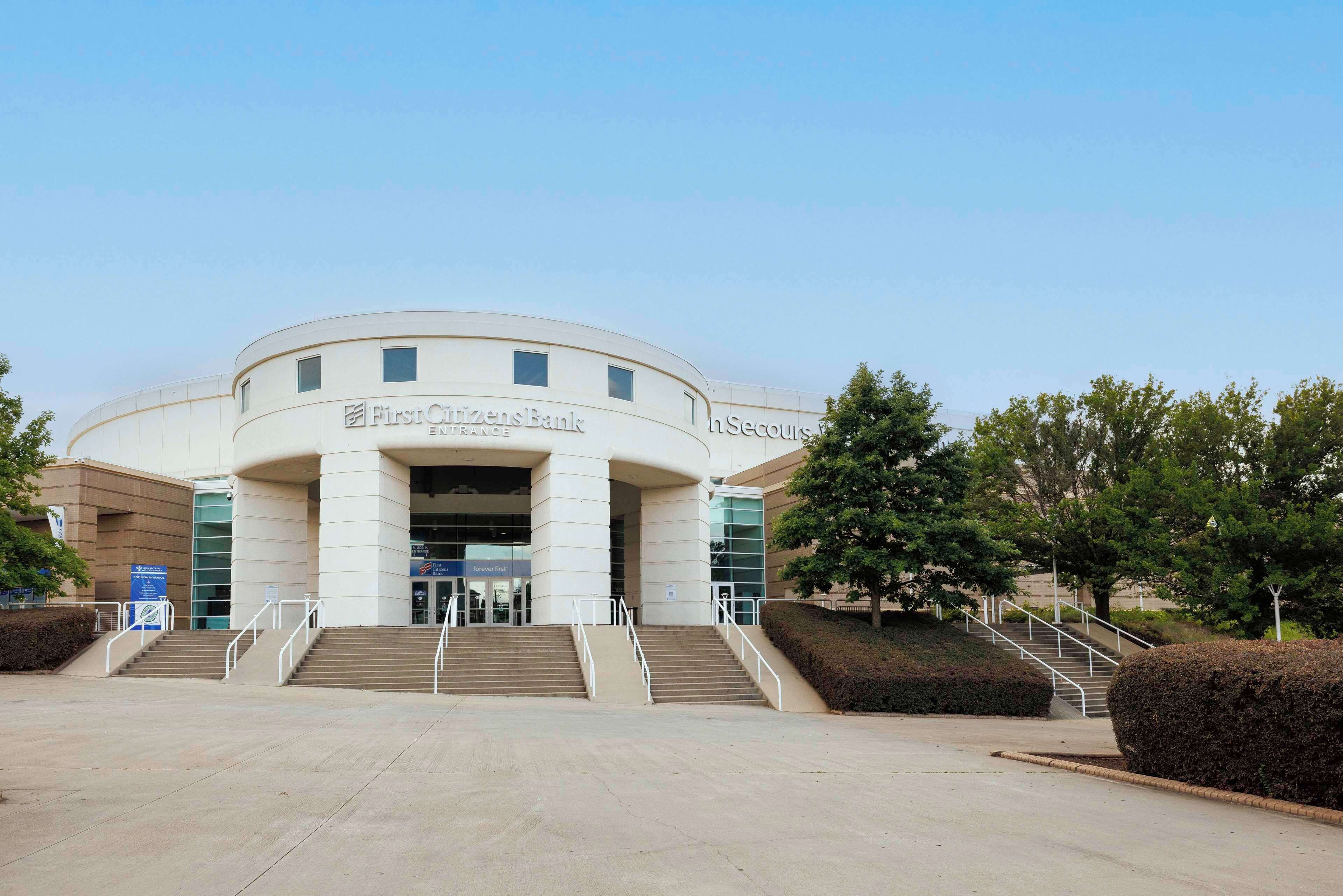 A building with a curved facade, labeled "First Citizens Bank" and "The Secours." It features a set of stairs leading to the entrance, surrounded by landscaped areas and trees.