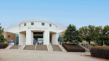 A building with a curved facade, labeled "First Citizens Bank" and "The Secours." It features a set of stairs leading to the entrance, surrounded by landscaped areas and trees.