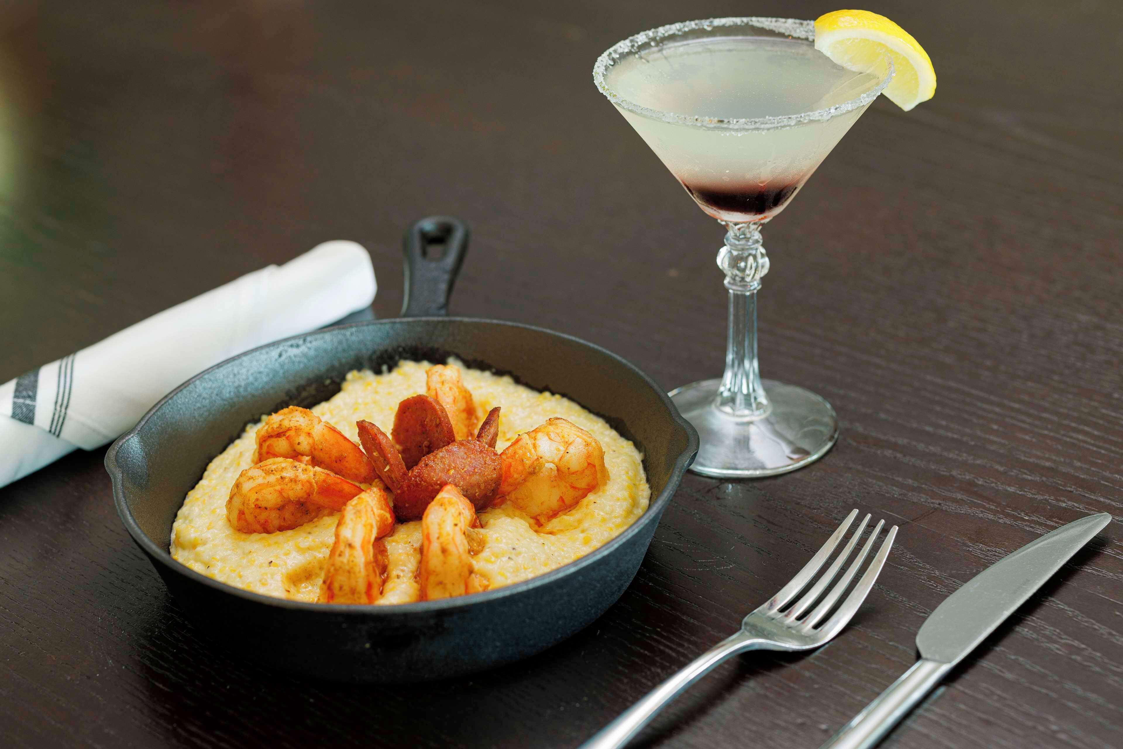 A skillet with shrimp and polenta, a martini glass with a lemon garnish, and cutlery on a wooden table.