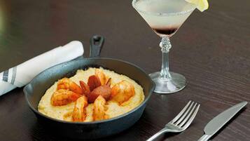 A skillet with shrimp and polenta, a martini glass with a lemon garnish, and cutlery on a wooden table.
