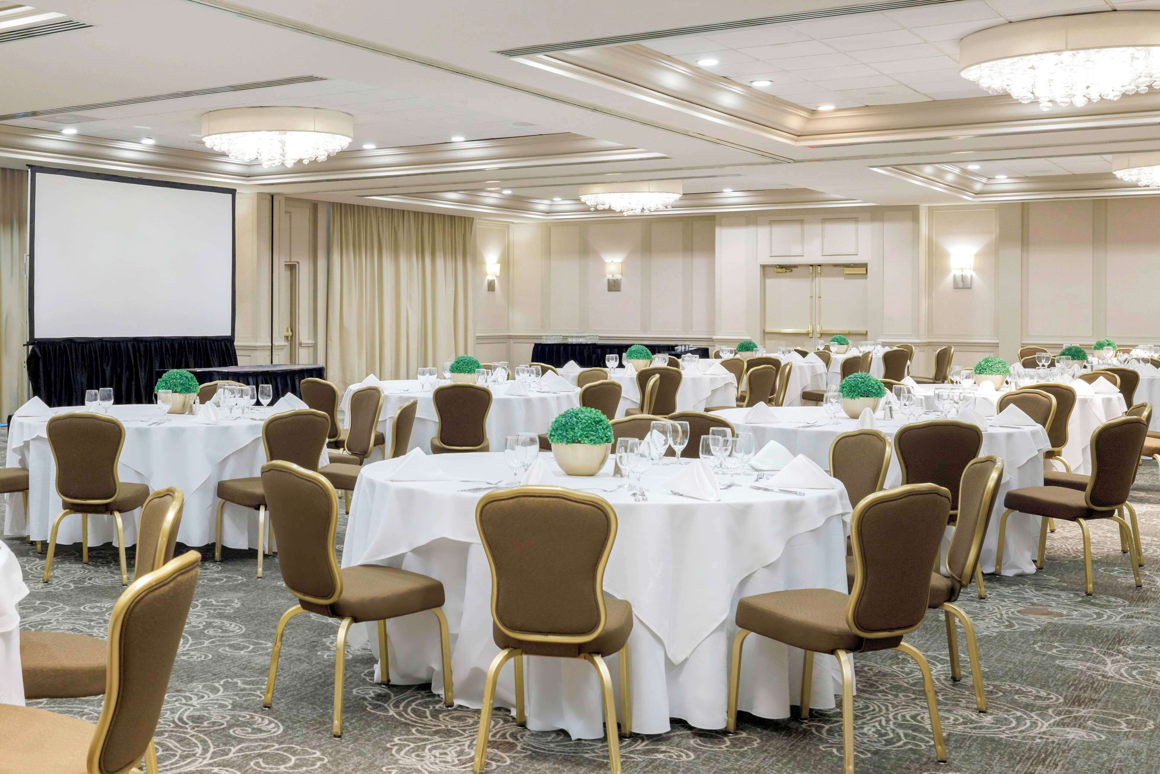 Un salón de banquetes con mesas redondas, sillas marrones y manteles blancos.