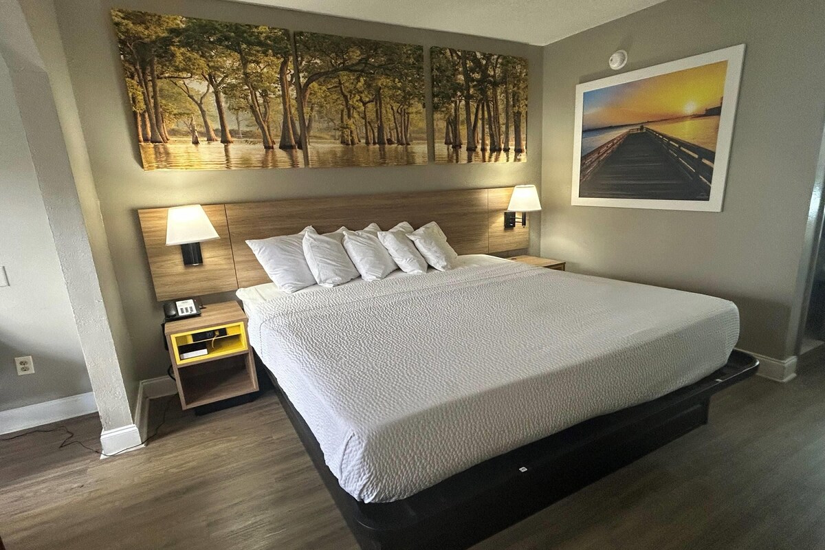 A neatly made bed with a white comforter, a wooden headboard, two bedside lamps, a nightstand with a lamp, and a large painting of a sunset over water on the wall.