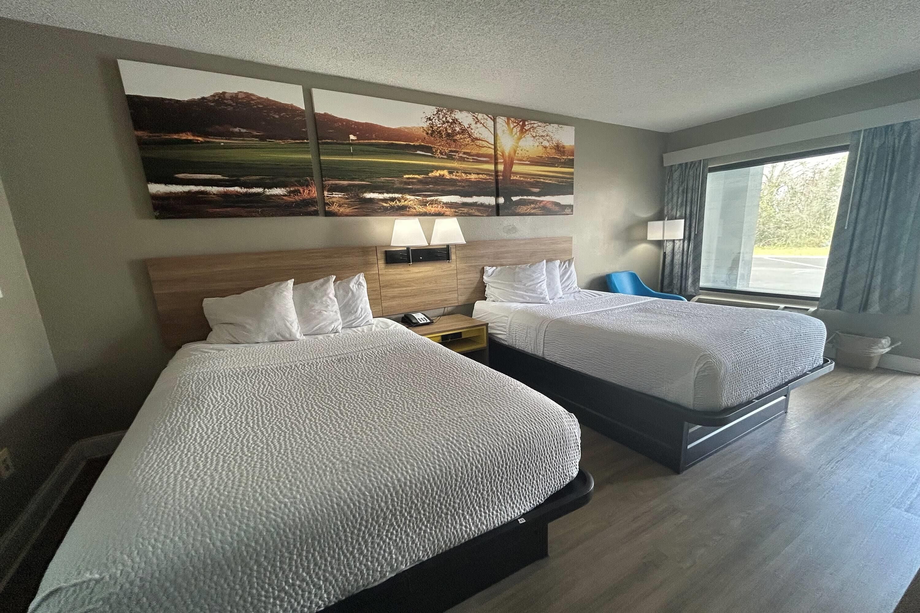 Habitación de hotel con dos camas, un cuadro grande sobre ellas, una mesita de noche con una lámpara y una ventana con cortinas.