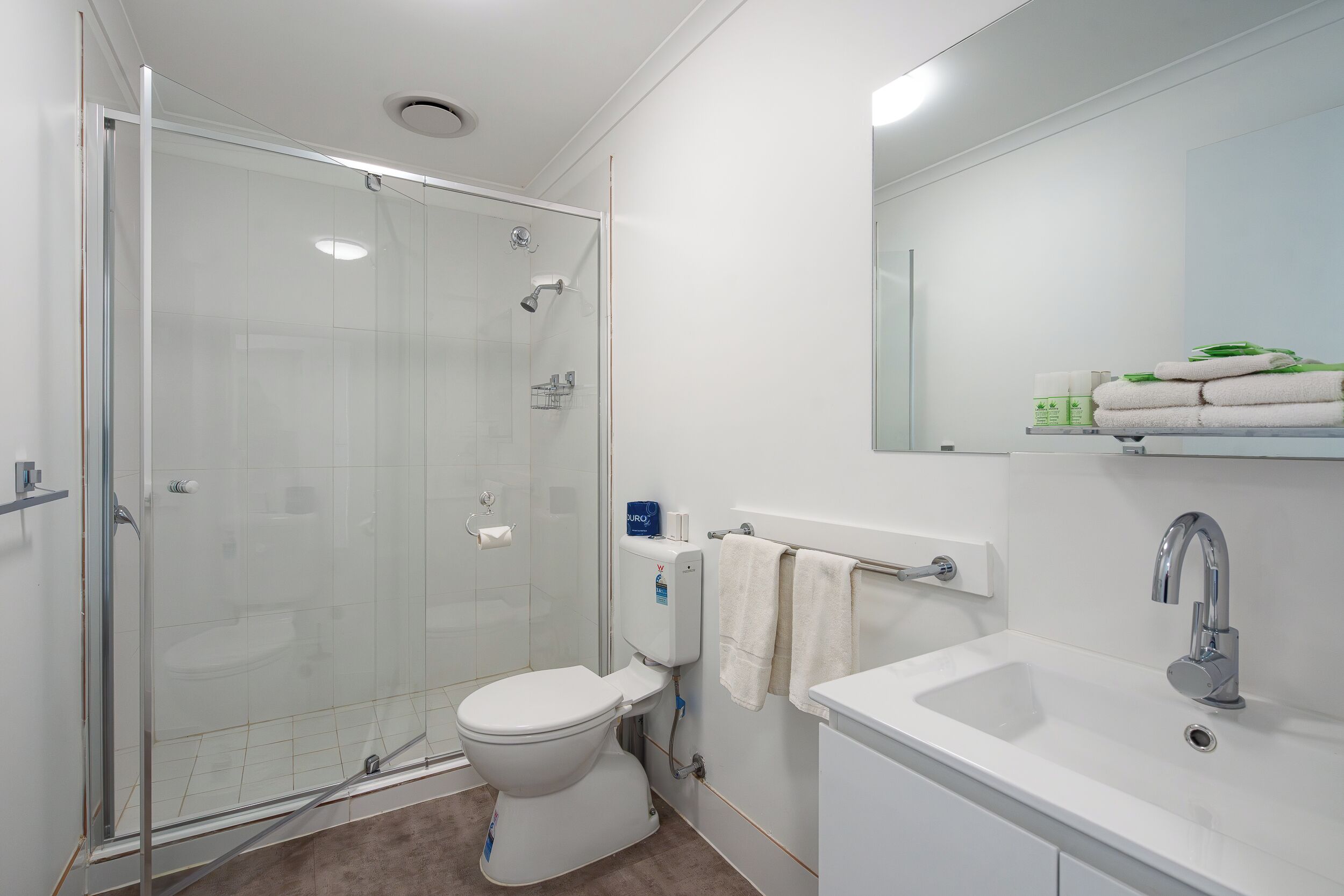 A modern bathroom with a glass-enclosed shower, a white toilet, a sink with a mirror, and neatly folded towels on a shelf.