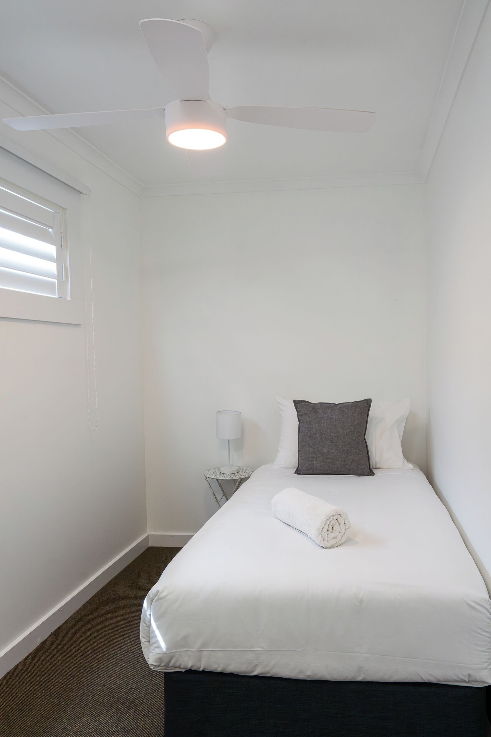 A single bed with white bedding, a grey pillow, a white lamp, and a ceiling fan in a room with white walls and a carpeted floor.