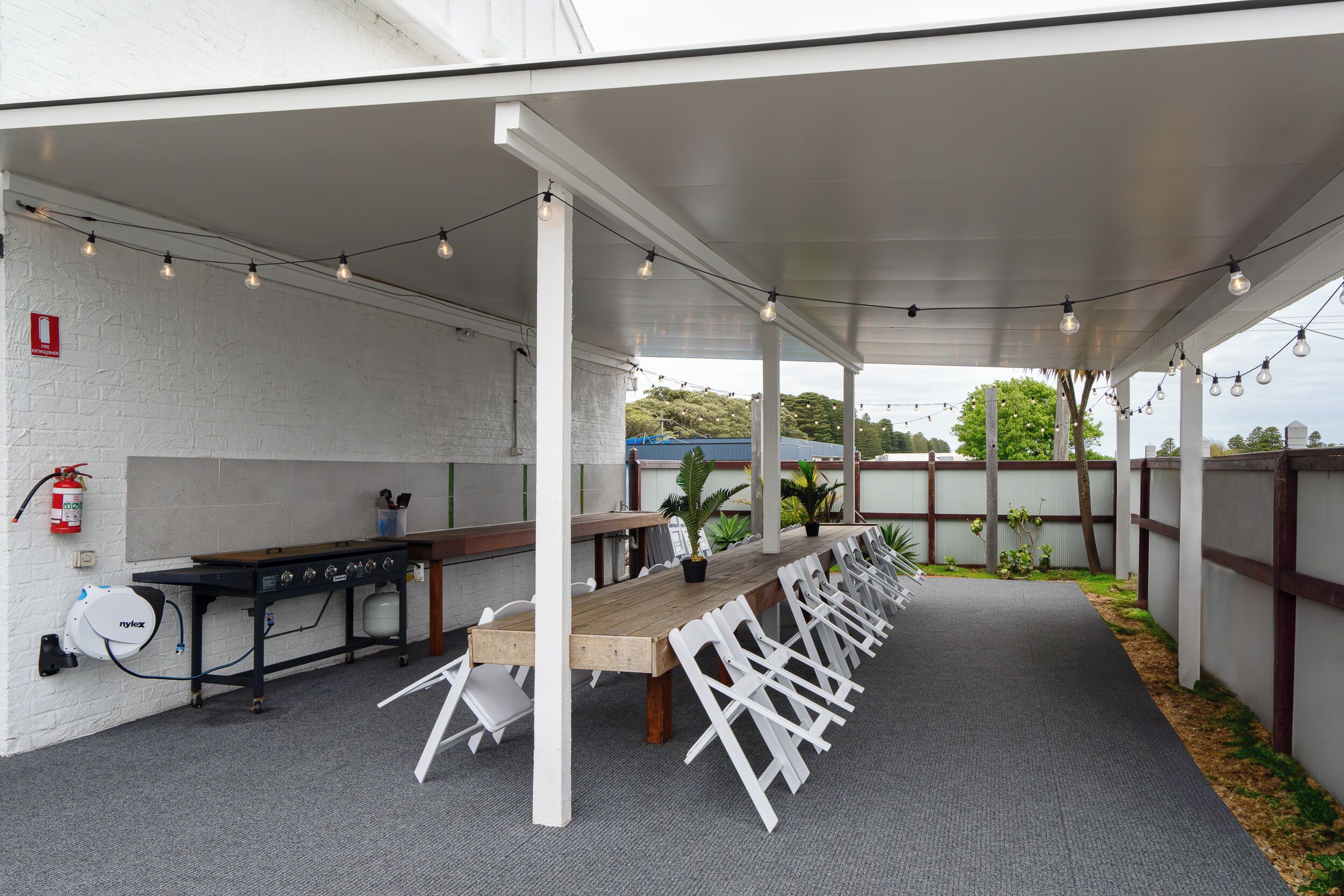 An outdoor covered patio with a long wooden table and white chairs, a grill, and string lights.