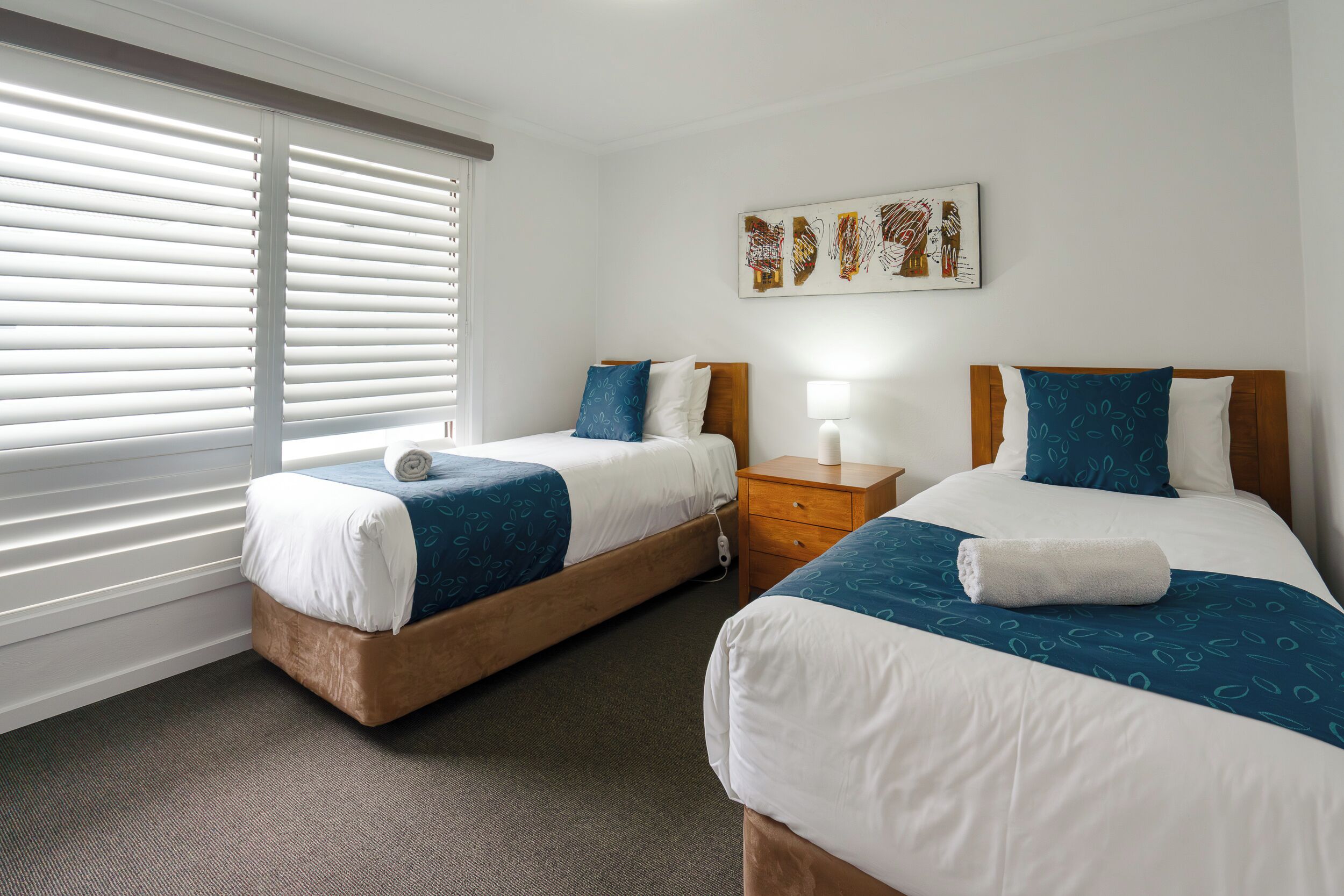 A hotel room with two beds, a nightstand, a lamp, and a window with blinds.