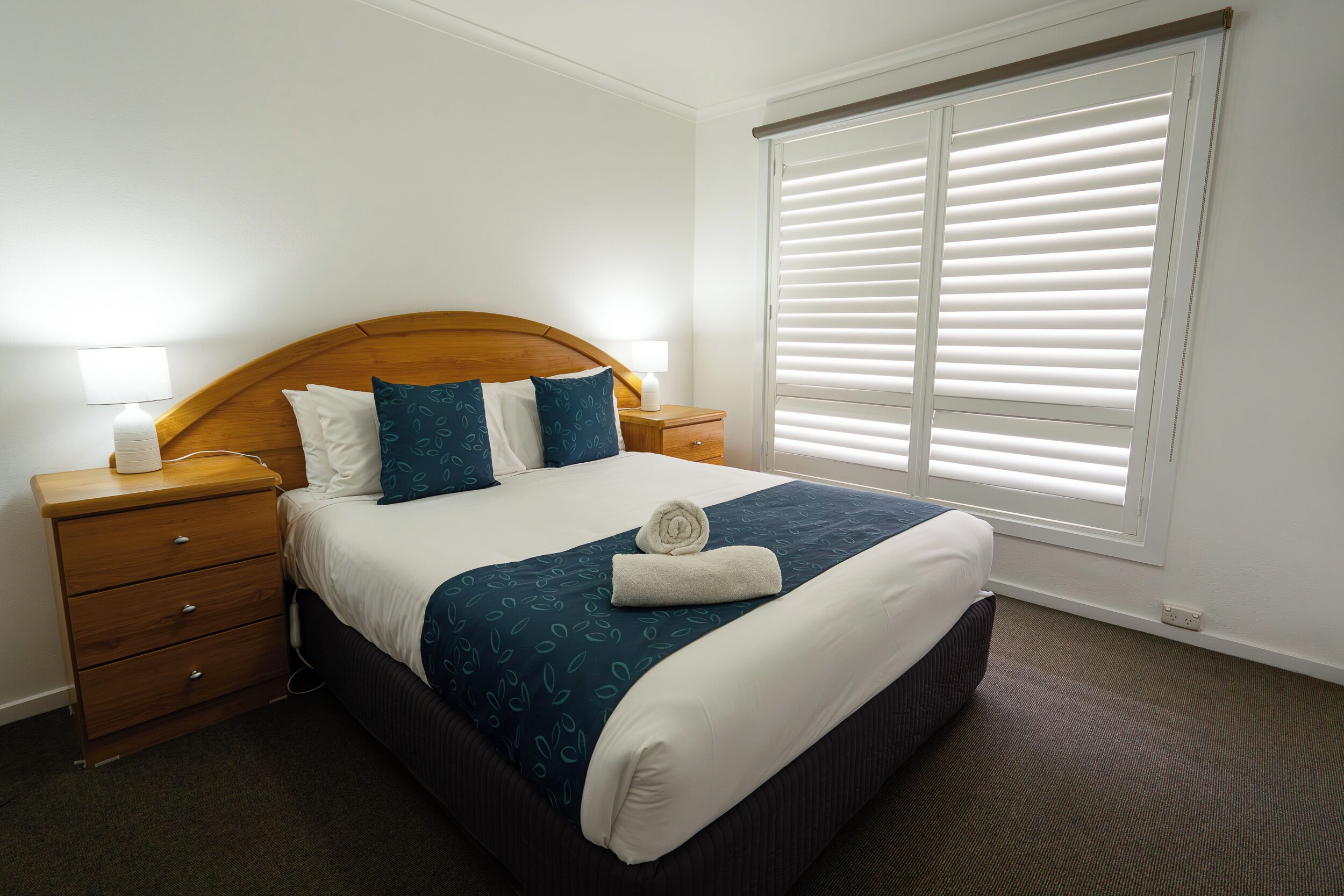 Cama bem arrumada com colcha azul e branca estampada, dois travesseiros, cabeceira de madeira e mesa de cabeceira com abajur.