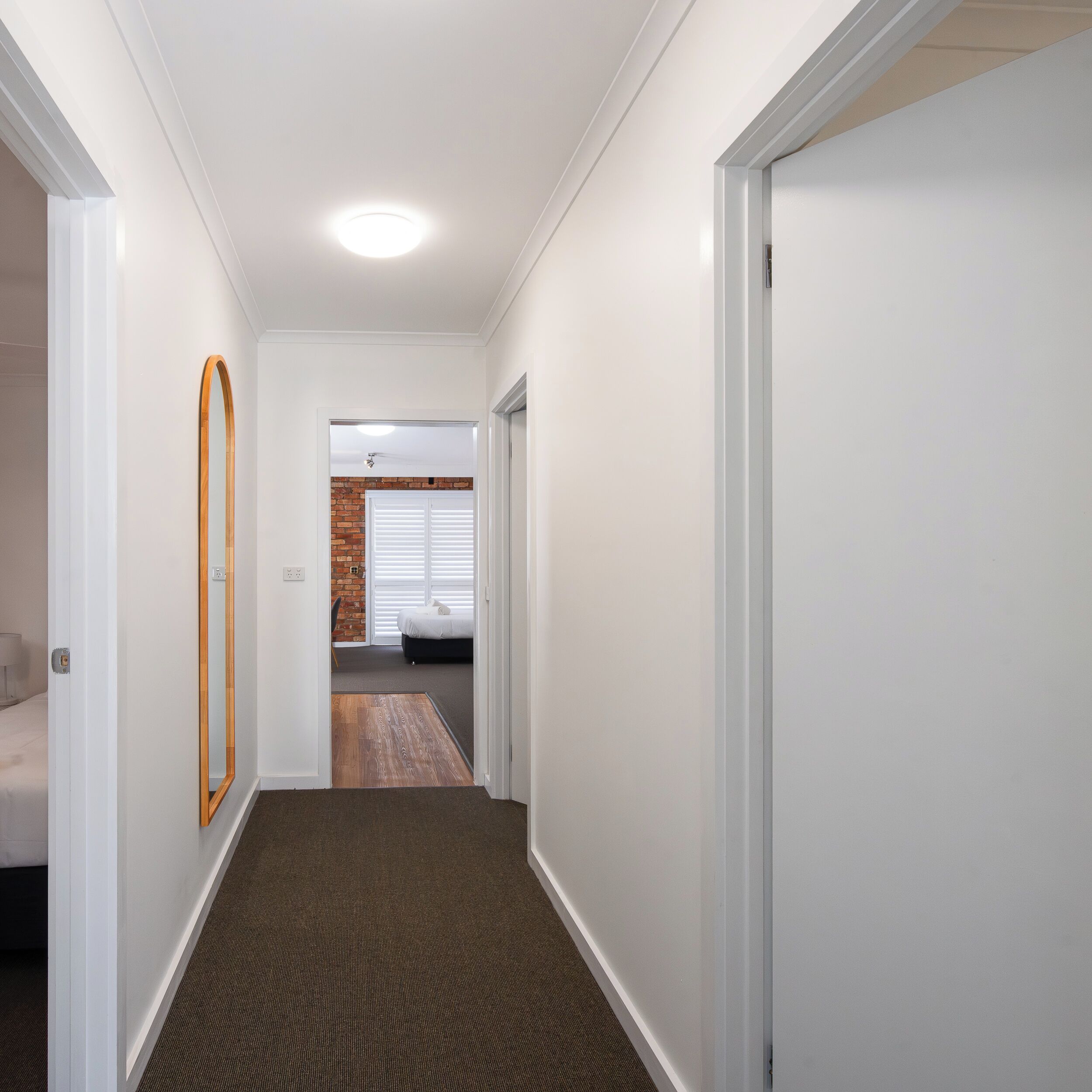 Un pasillo con paredes blancas, suelo de madera y una puerta que da a un dormitorio con pared de ladrillo y una cama.