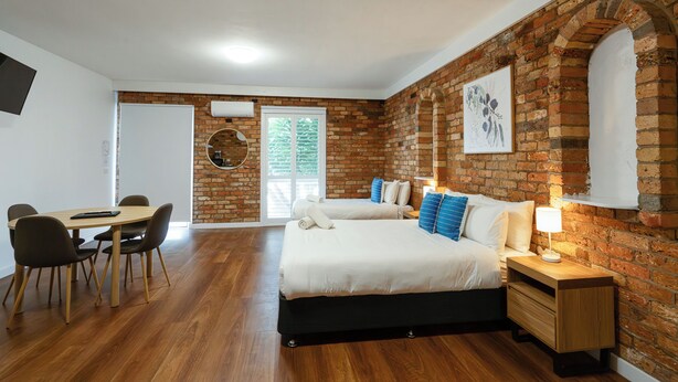 A bedroom with a brick wall, a bed with white bedding, a bedside table with a lamp, and a dining area with chairs and a table.