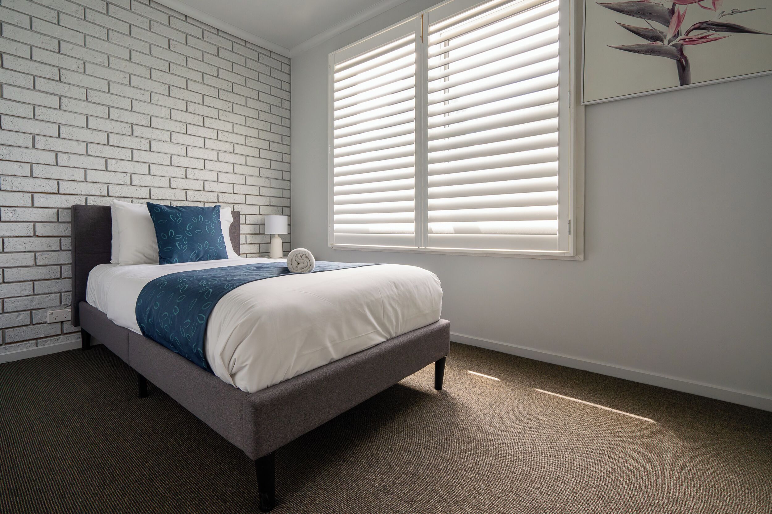 A bedroom with a bed, a brick wall, a window with blinds, and a painting on the wall.