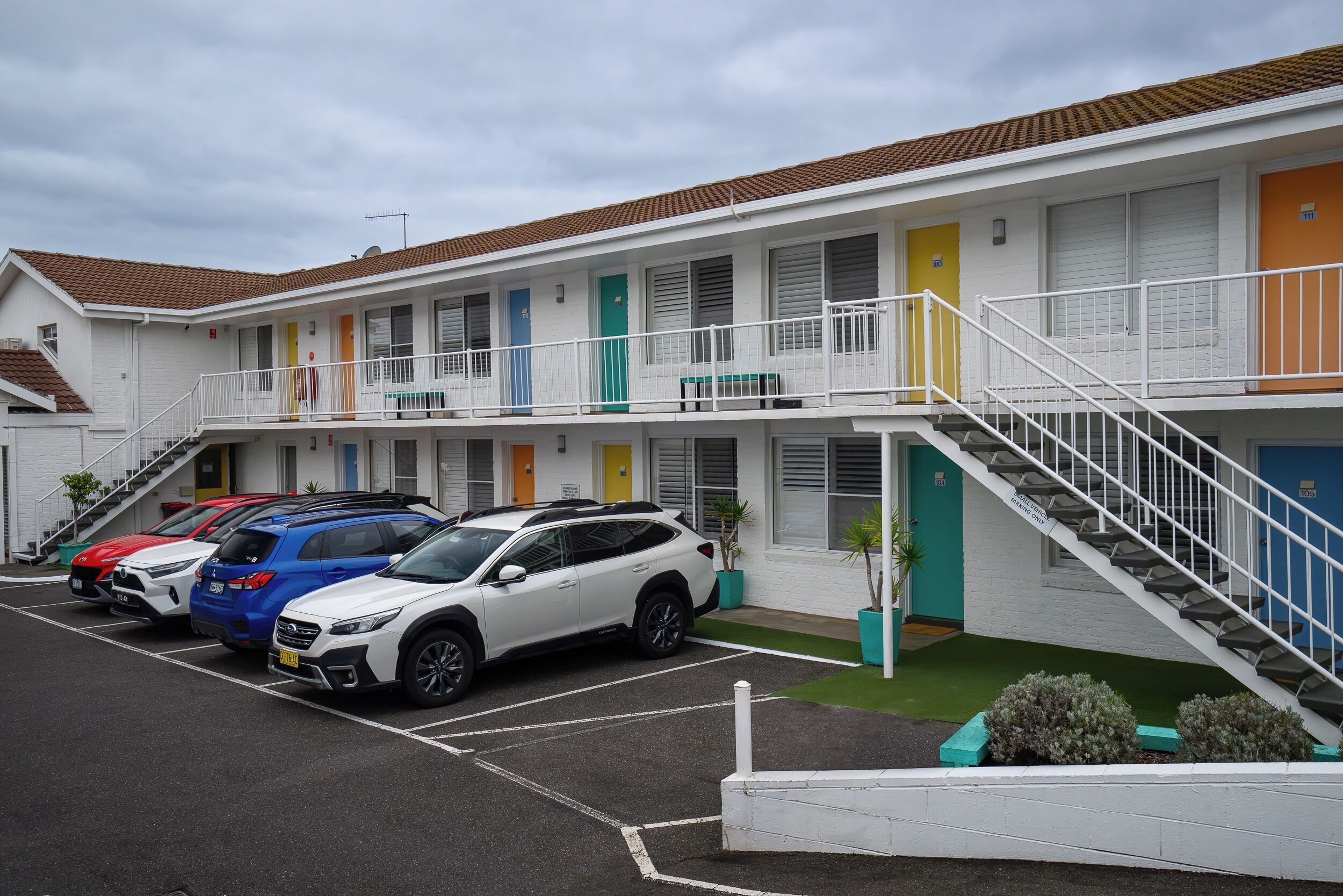 A multi-story motel with a parking lot, a staircase, and a row of colorful doors.