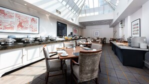 Een ruime eetzaal van een hotel met een hoge plafond en dakraam, met een lange balie met food stations, meerdere eettafels en comfortabele zitplaatsen.